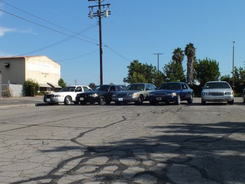 2003 Ford Crown Victoria - Five Panther-platform sedans parked at an angle on asphalt beside a white industrial warehouse.