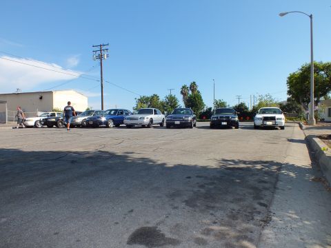 2003 Ford Crown Victoria - Cars sit angled across a wide lot while a few people gather at the left, building beyond.