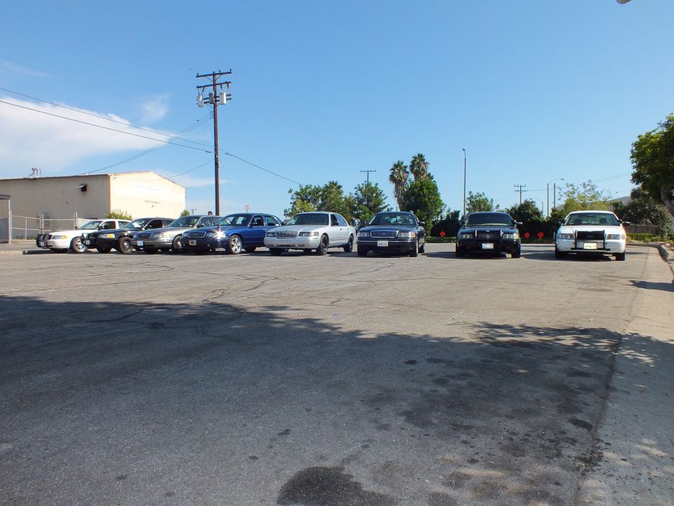 Eight sedans parked in a row along a warehouse wall in a paved lot