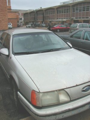 1997 Ford Taurus - Weathered silver first-generation Ford Taurus sedan parked in a lot, shot from the front corner showing heavily oxidized and peeling paint…
