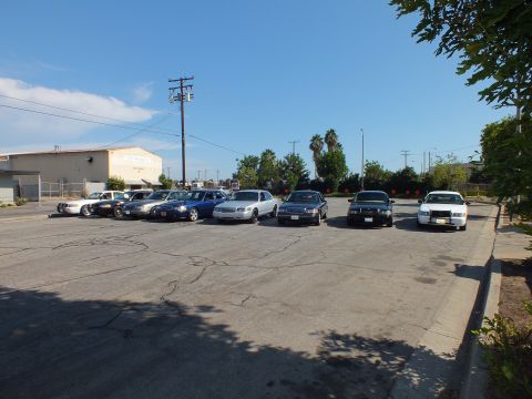 2003 Ford Crown Victoria - Mixed sedans angle across a sunlit lot, palm trees and a low warehouse in the background.