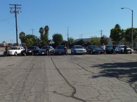 2003 Ford Crown Victoria - Owners pose alongside their full-size sedans lined up across a sunlit industrial lot.