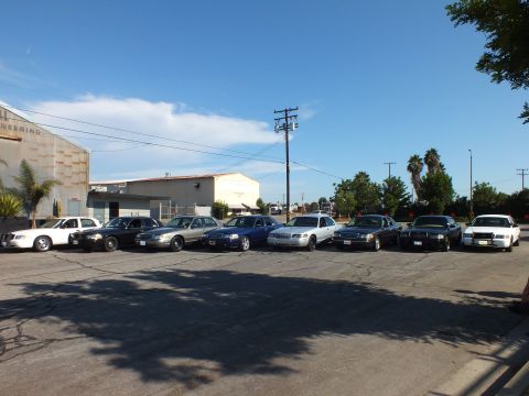 2003 Ford Crown Victoria - Sedans line up front-on beside a warehouse with faded Sheet Metal Engineering lettering.