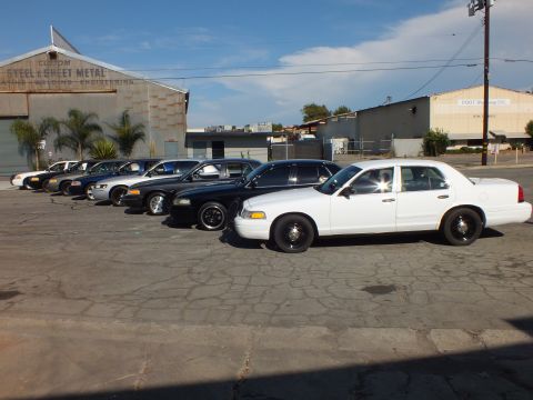 2003 Ford Crown Victoria - white Crown Victoria heads a diagonal line of sedans by a Custom Steel and Sheet Metal shop.