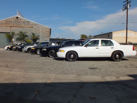 2003 Ford Crown Victoria - white Crown Victoria fronts a row of black and silver sedans by a sheet metal warehouse.