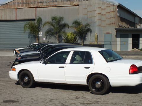 2003 Ford Crown Victoria - white Crown Victoria sits in full profile ahead of black sedans by a corrugated warehouse.