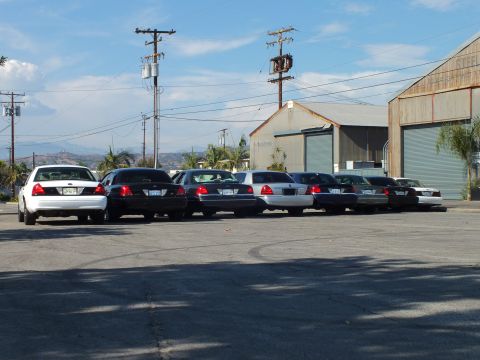 2003 Ford Crown Victoria - Several full-size sedans lined up rear-to-camera near metal warehouses, power poles and distant hills