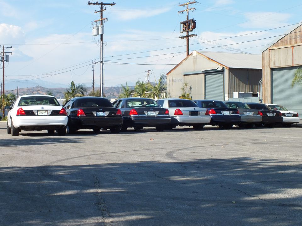 Rear view of seven parked sedans in industrial lot with warehouse behind