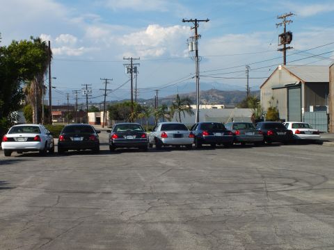 2003 Ford Crown Victoria - Long row of dark and silver sedans seen from rear with mountains and warehouse in distance