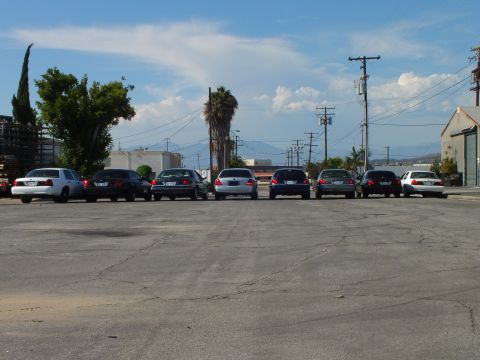 2003 Ford Crown Victoria - Lineup of full-size sedans on weathered asphalt, trees and warehouse on the horizon
