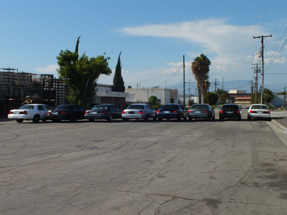 Eight sedans parked across asphalt lot with industrial buildings behind