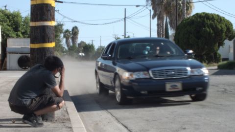 2003 Ford Crown Victoria - Photographer kneels at curb shooting a dark sedan rolling past on a sunny street, palms behind