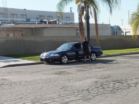 2003 Ford Crown Victoria - Lowered dark sedan on cracked street, man beside open driver door, block wall and palm behind