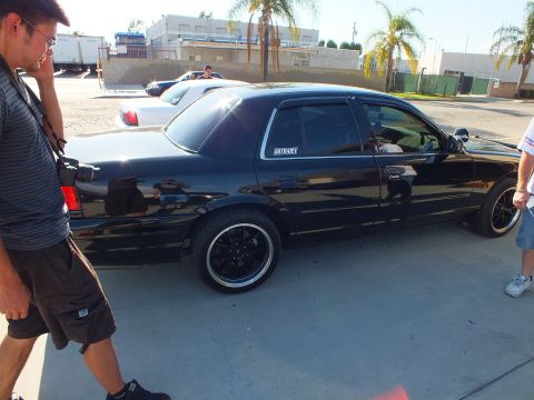 2003 Ford Crown Victoria - Side view of glossy black full-size sedan on dark wheels, two men nearby and white car behind