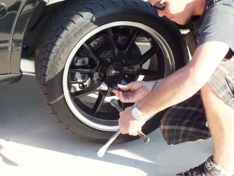 2003 Ford Crown Victoria - Kneeling next to a black sedan, hands work a socket on the SVT-capped black multi-spoke wheel