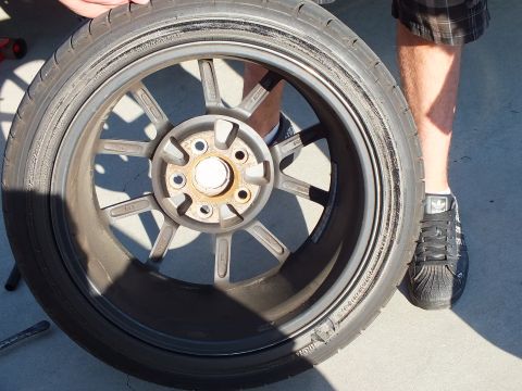 2003 Ford Crown Victoria - dismounted silver multi-spoke wheel showing a surface-rusted hub sits on the ground by a leg