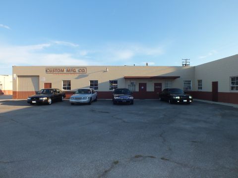 2003 Ford Crown Victoria - Row of four full-size sedans, headlights on, parked before the Custom Mfg. Co. building