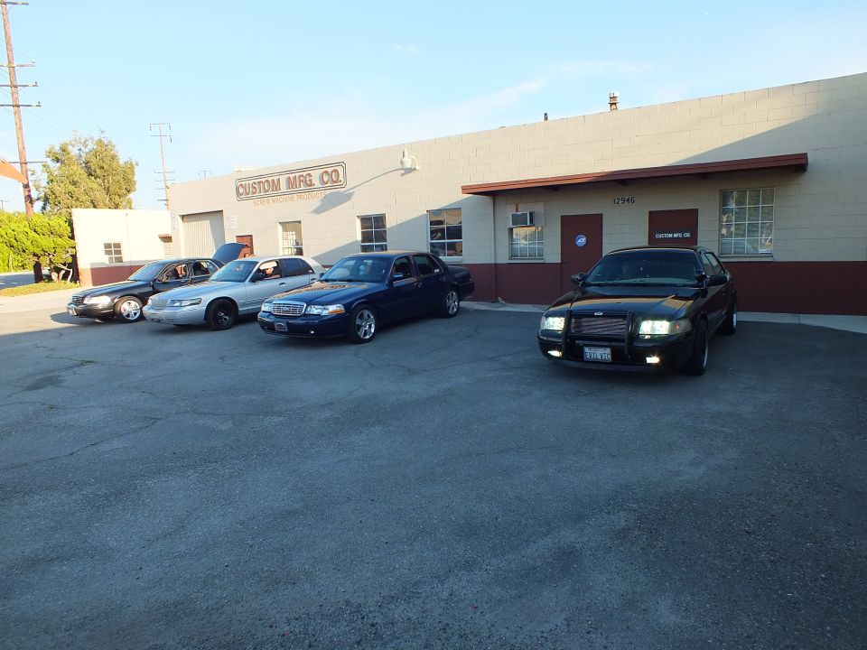 Black sedan with headlights on angled in lot beside three sedans at Custom Mfg Co