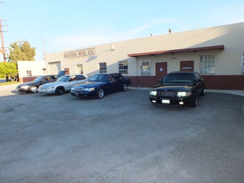 2003 Ford Crown Victoria - Black sedan, lights lit, parked among silver and blue full-size sedans outside an industrial shop