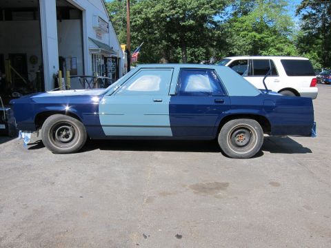 1991 Ford LTD Crown Victoria - A two-tone blue sedan, possibly undergoing repairs or a paint job, is parked outside a commercial garage with a white SUV in the background.