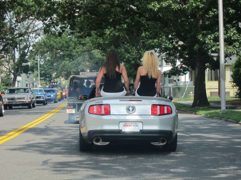1991 Ford LTD Crown Victoria - Rear view of a silver Mustang convertible carrying two women in a parade on a tree-lined street with vintage trucks behind.