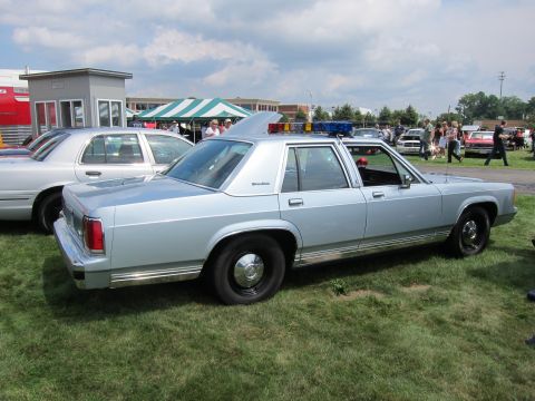 1991 Ford LTD Crown Victoria - light-blue full-size Ford sedan with a roof light bar parked on grass at an outdoor car show with crowds and tents behind.