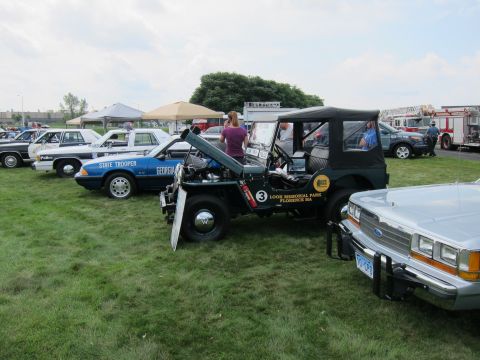 1991 Ford LTD Crown Victoria - vintage soft-top police Jeep with its hood raised is displayed among State Trooper and patrol cars on a grassy show field.