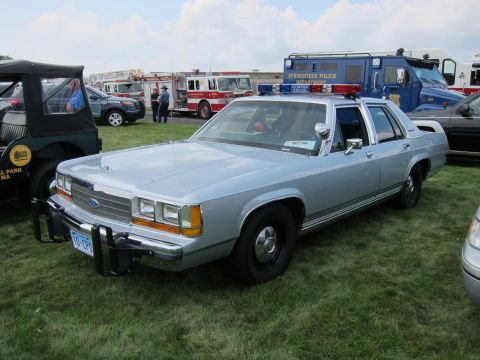 1991 Ford LTD Crown Victoria - silver full-size Ford police sedan with push bar and light bar parked on grass with fire apparatus in the background.