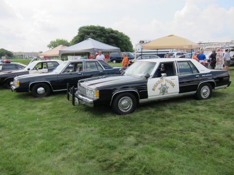 1991 Ford LTD Crown Victoria - black-and-white 'Highway Patrol' Ford sedan with a push bar parked on grass beside other cruisers at a crowded car show.
