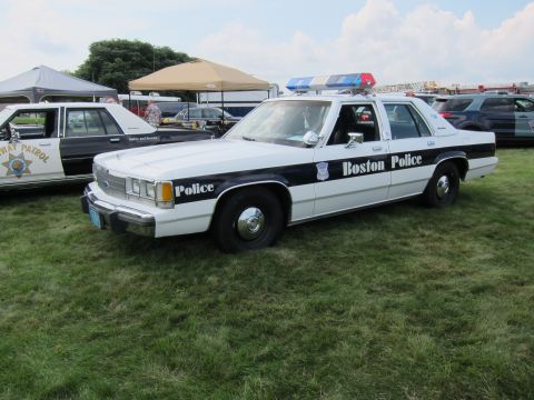 1991 Ford LTD Crown Victoria - black-and-white Boston Police Ford sedan with a roof light bar parked on grass near tents and other cruisers at a car show.