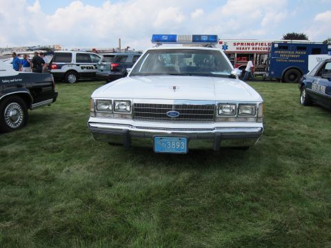 1991 Ford LTD Crown Victoria - Head-on view of a white Ford police cruiser with a blue light bar and Massachusetts '3893' plate on grass near rescue trucks.