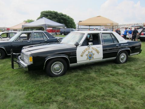 1991 Ford LTD Crown Victoria - black-and-white 'Highway Patrol' Ford sedan with a star door shield and push bar parked on grass under tents at a car show.