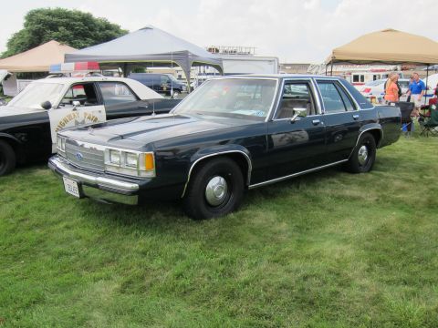 1991 Ford LTD Crown Victoria - dark blue Ford four-door sedan with a Highway Patrol door emblem sits on grass beside other vehicles under show tents.