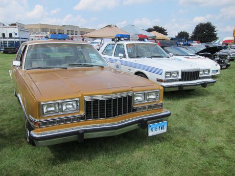 1991 Ford LTD Crown Victoria - gold full-size four-door sedan with stacked headlights sits front-on beside white police cars at a grass show.