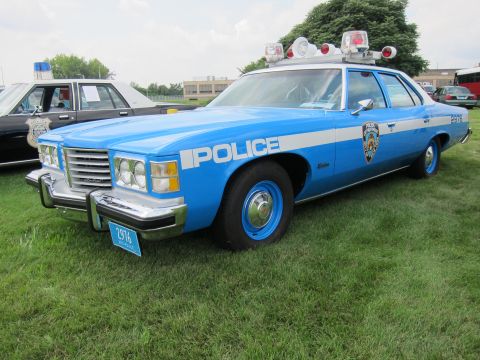 1991 Ford LTD Crown Victoria - Front-quarter view of a light-blue Pontiac police sedan with POLICE lettering and roof spotlights on a grass field.