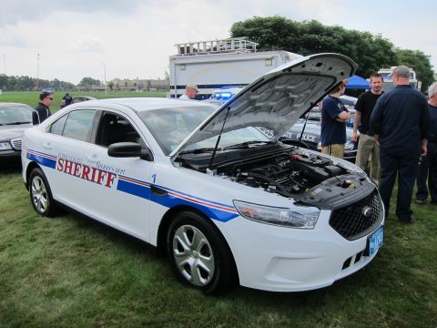 1991 Ford LTD Crown Victoria - white Ford Taurus Sheriff cruiser numbered 1 with its hood open to show the engine, displayed on grass as people stand nearby.