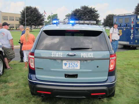 1991 Ford LTD Crown Victoria - rear of a blue Ford Explorer State Police SUV with blue roof lights active and a Massachusetts 1526 trooper plate, spectators alongside.