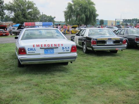 1991 Ford LTD Crown Victoria - Two vintage police sedans on grass: a white car lettered EMERGENCY CALL 9-1-1 beside a black HIGHWAY PATROL sedan with chrome bumpers.