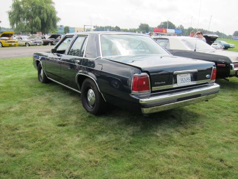 1991 Ford LTD Crown Victoria - plain dark-blue Ford Crown Victoria sedan with black steel wheels, viewed from the rear three-quarter on grass among other show cars.