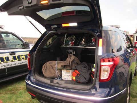 1991 Ford LTD Crown Victoria - Rear hatch open on a dark-blue Ford Explorer police SUV loaded with bags and gear, parked next to a New England University police vehicle.