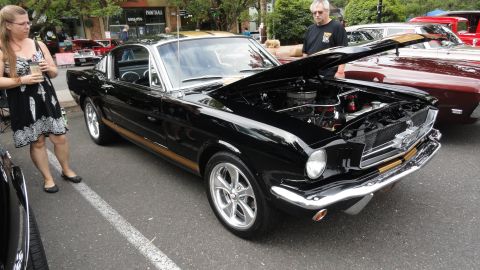 1964 Ford Fairlane - A black 1965 Ford Mustang fastback with a gold stripe and its hood open is displayed at an outdoor event.