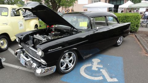 1964 Ford Fairlane - A black Chevrolet sedan from the mid-1950s is parked in a handicapped spot, its hood open to reveal the engine.