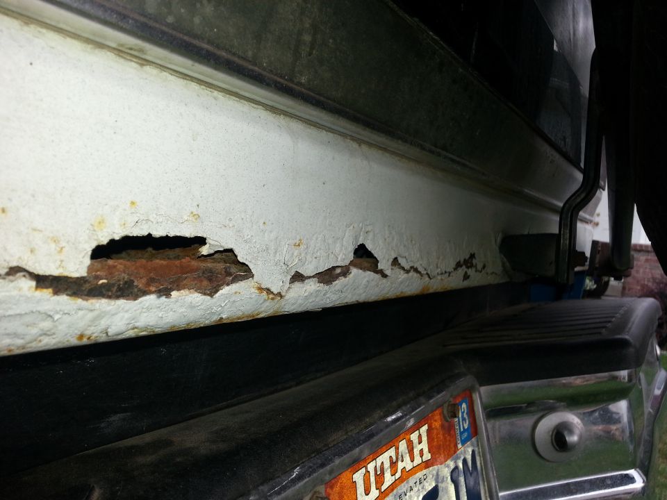 Close-up of severe rust damage on a white vehicle's rocker panel above the rear bumper. Utah license plate visible