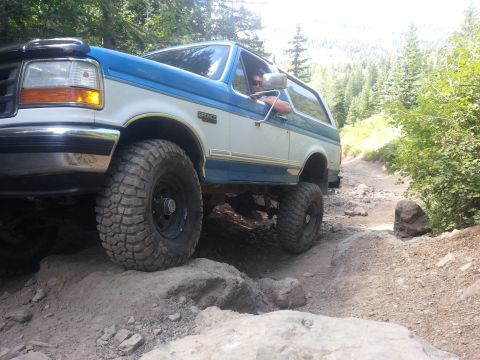 1994 Ford Bronco - A blue and white Ford Bronco navigates a rocky off-road trail with trees in the background.