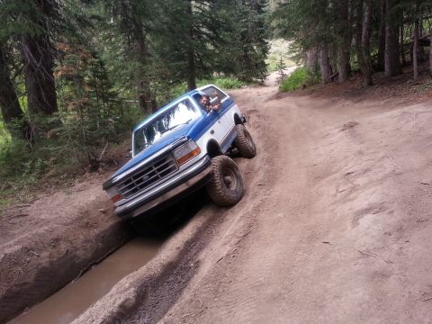1994 Ford Bronco - A blue and white Ford Bronco drives through a challenging muddy terrain on a wooded trail.