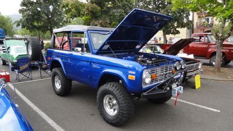 1964 Ford Fairlane - A bright blue Ford Bronco is displayed with its hood open, revealing the engine bay, alongside other vehicles.