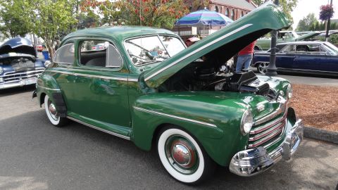 1964 Ford Fairlane - A green coupe with its hood open, revealing the engine, sits on display with whitewall tires.