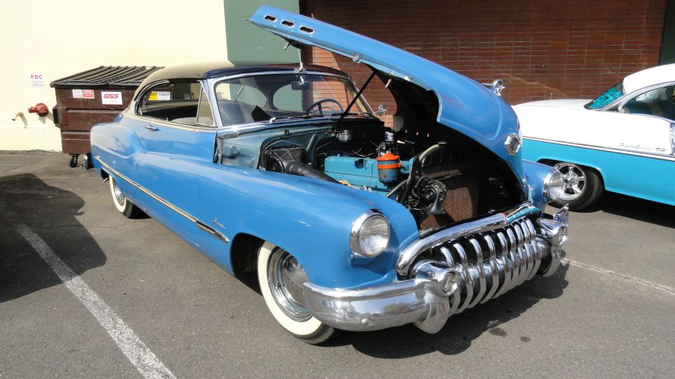 Blue classic car with hood open, showing engine. Another blue and white classic car is parked next to it
