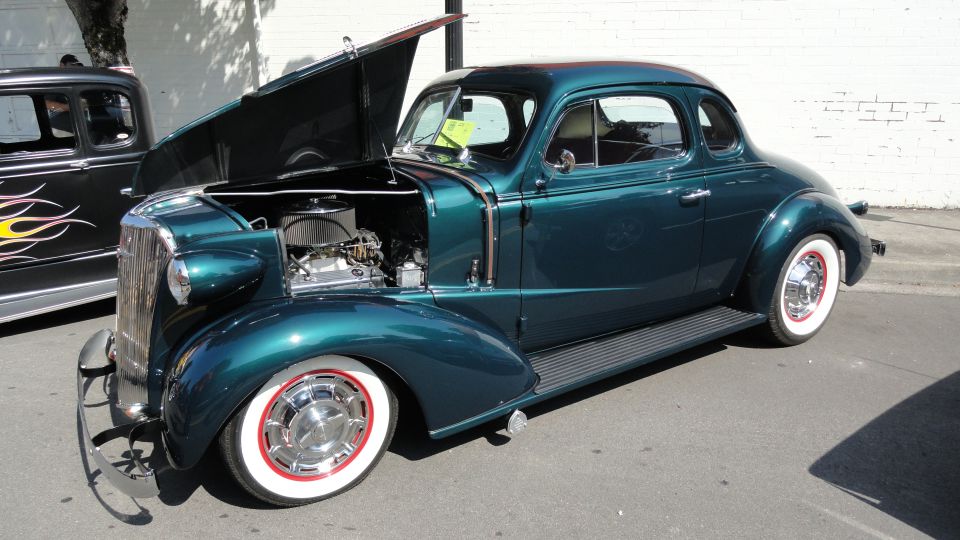 Dark teal coupe, hood up showing engine, white wall tires with red trim. Another car with flame decals is next to it