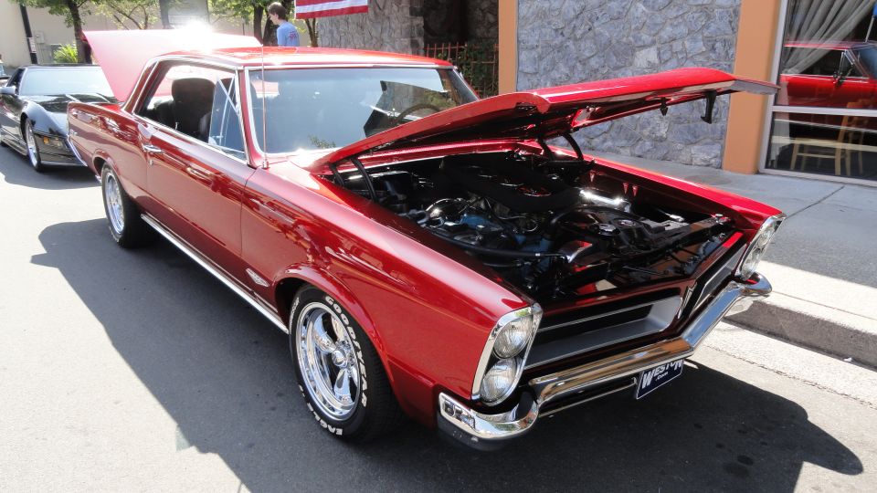 Red 1965 Pontiac Tempest coupe with hood open, showing engine. Another car is parked behind it